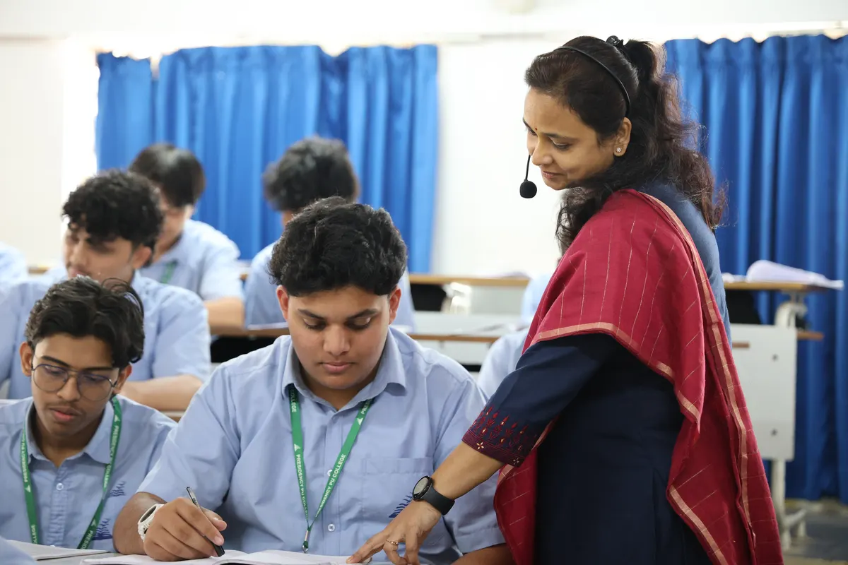 A teacher in a red saree with a headset microphone bends over a student's notebook in class, with blue curtains behind.
