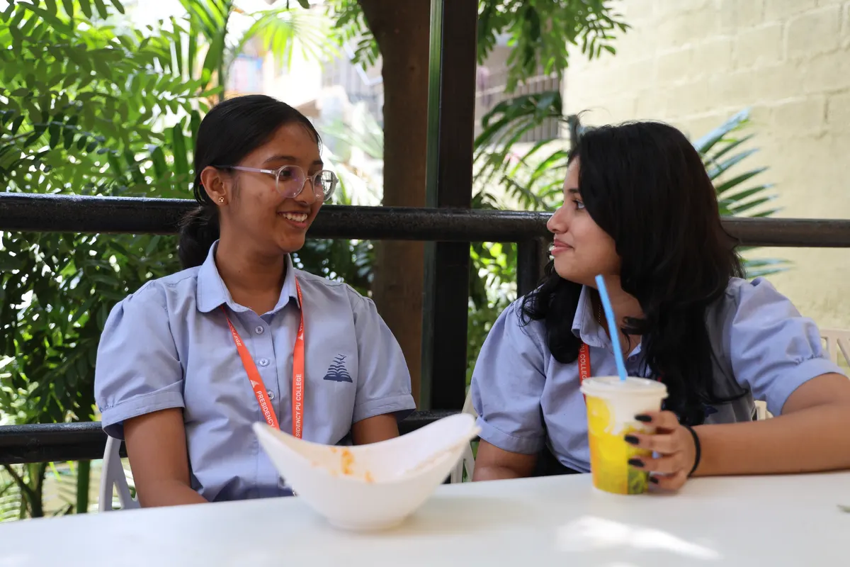Two students talking over drinks at an outdoor table under the trees.