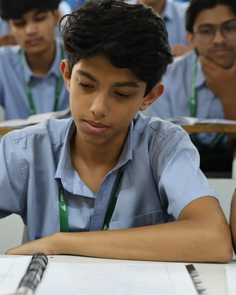 A Presidency PU student writing notes in class, with classmates seated behind him.